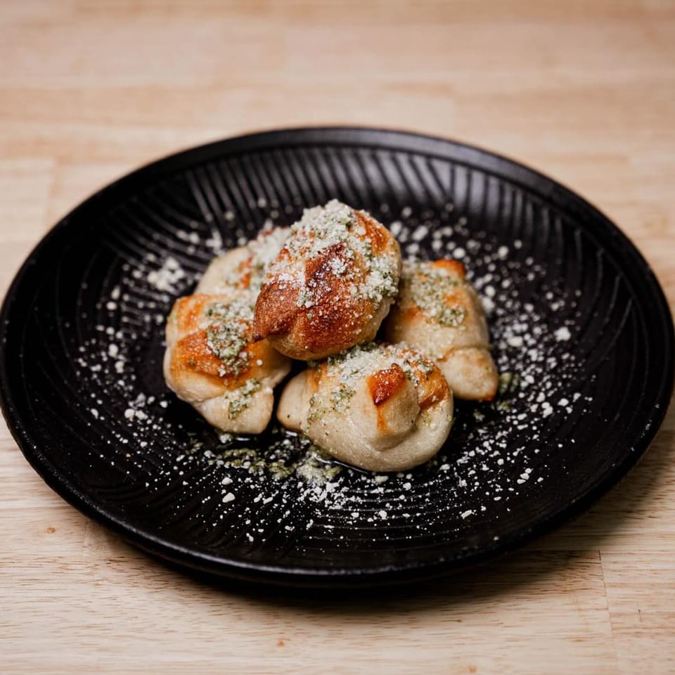Four Rocco's Pizza garlic knots, dusted with parmesan cheese and herbs, on a black plate.
