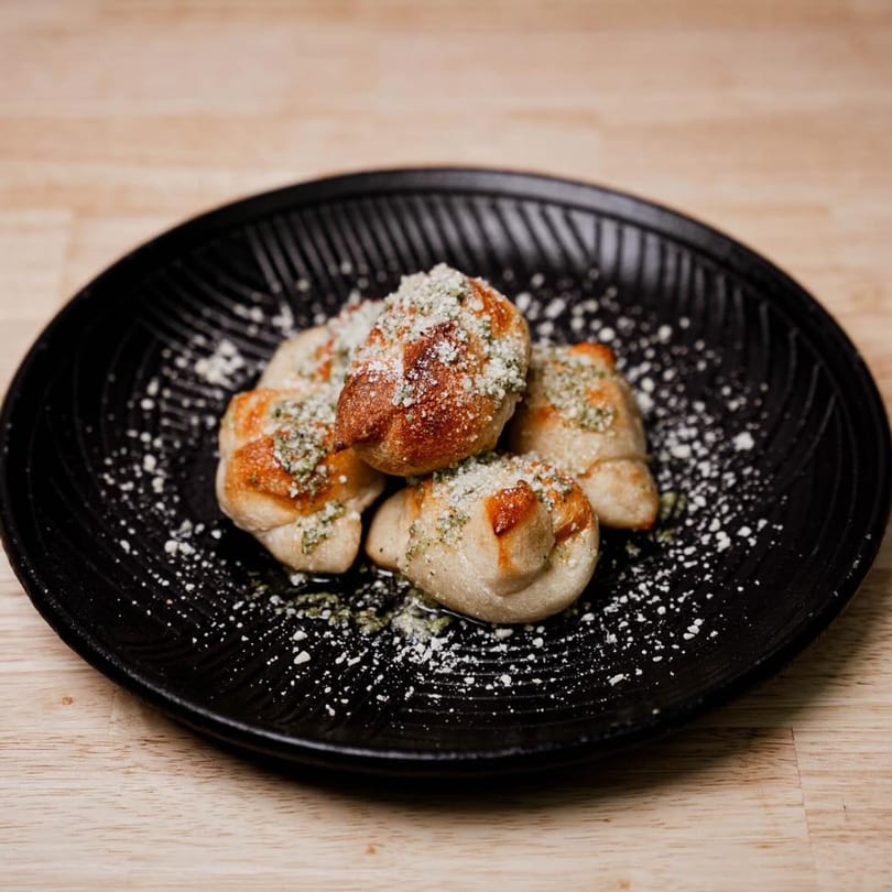 Four Rocco's Pizza garlic knots, dusted with parmesan cheese and herbs, on a black plate.
