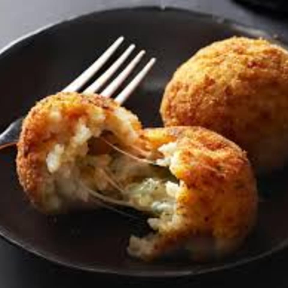 Two golden-brown arancini on a dark plate, one broken open with melted cheese stretching out.